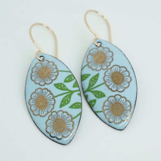 A pair of marquis-shaped drop earrings with a light blue background, decorated with gold flowers on green stems, hanging from gold hooks, against a white background