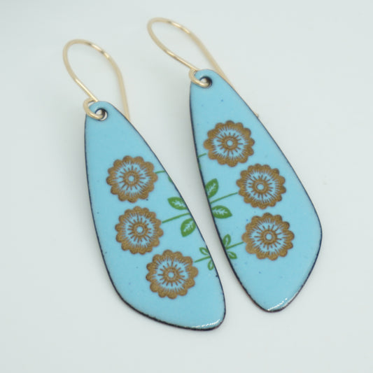 A pair of teardrop drop earrings with a light blue background, decorated with gold flowers on green stems, hanging from gold hooks, against a white background