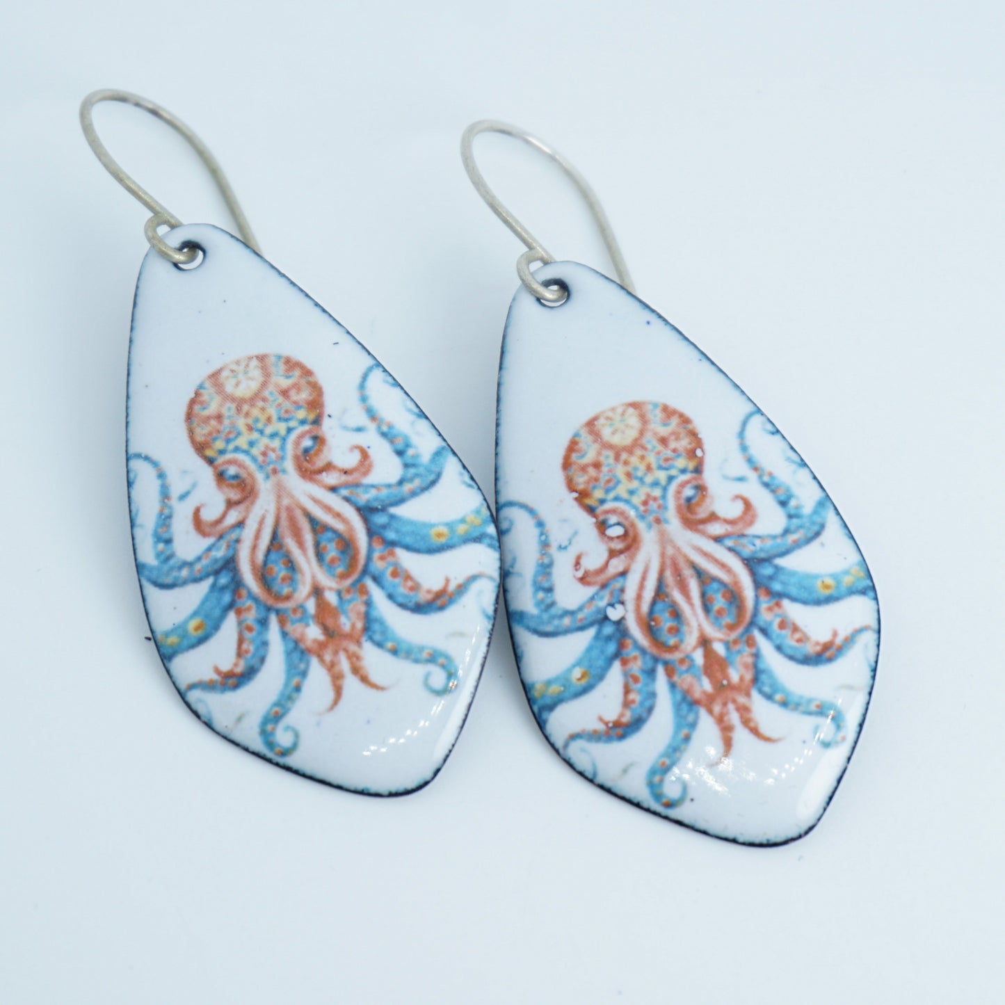 A pair of teardrop-shaped dangling earrings with silver fishhook wires. The glossy white enamel faces feature an octopus design in coral orange and light blue