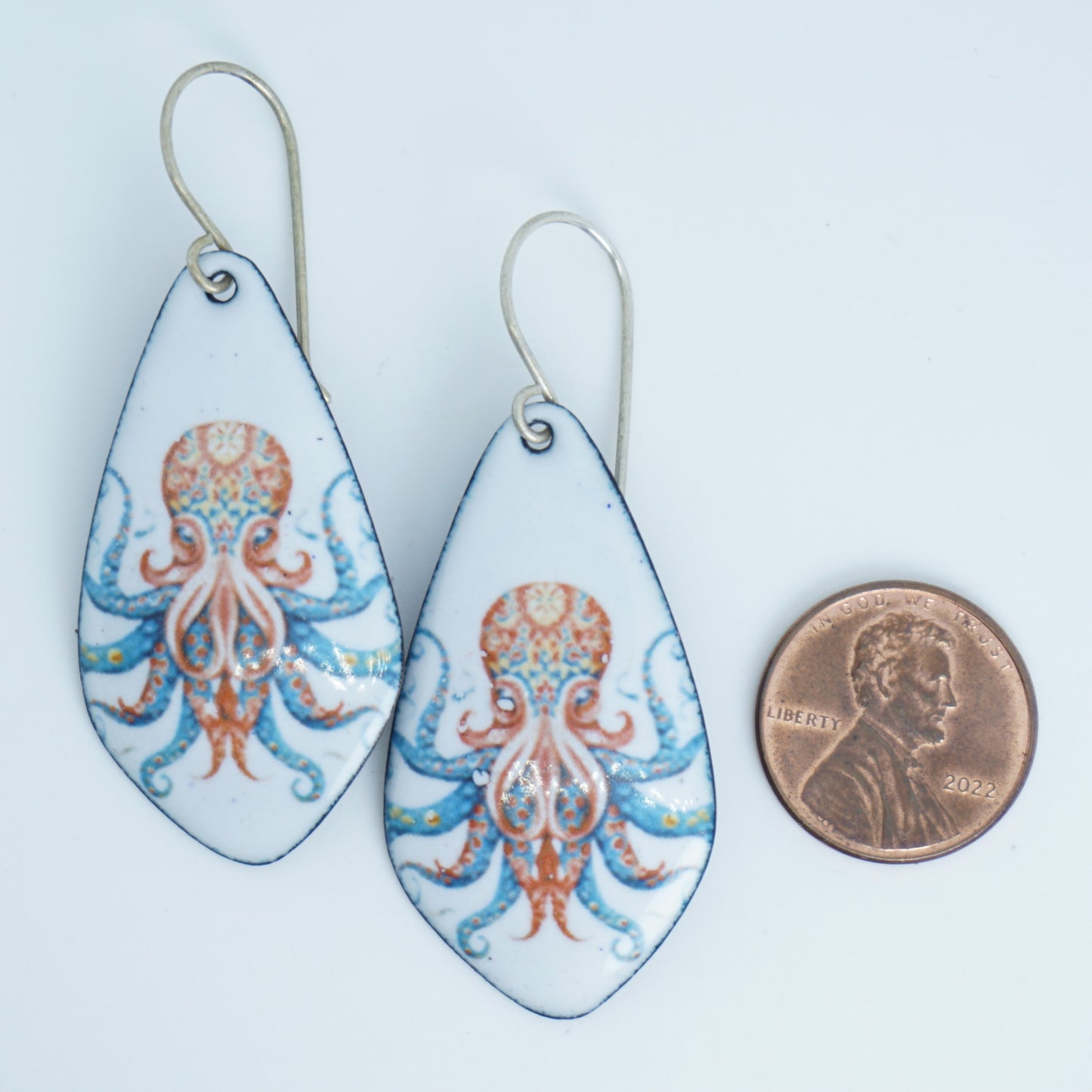 A pair of teardrop-shaped dangling earrings with a white background and an octopus design in blue and orange, shown next to a penny for scale on a white background.