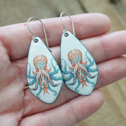 A pair of teardrop-shaped earrings with octopus illustrations, held in a person's hand. The earrings have a white background with a detailed octopus design in orange and blue, and are suspended from silver-tone hooks.