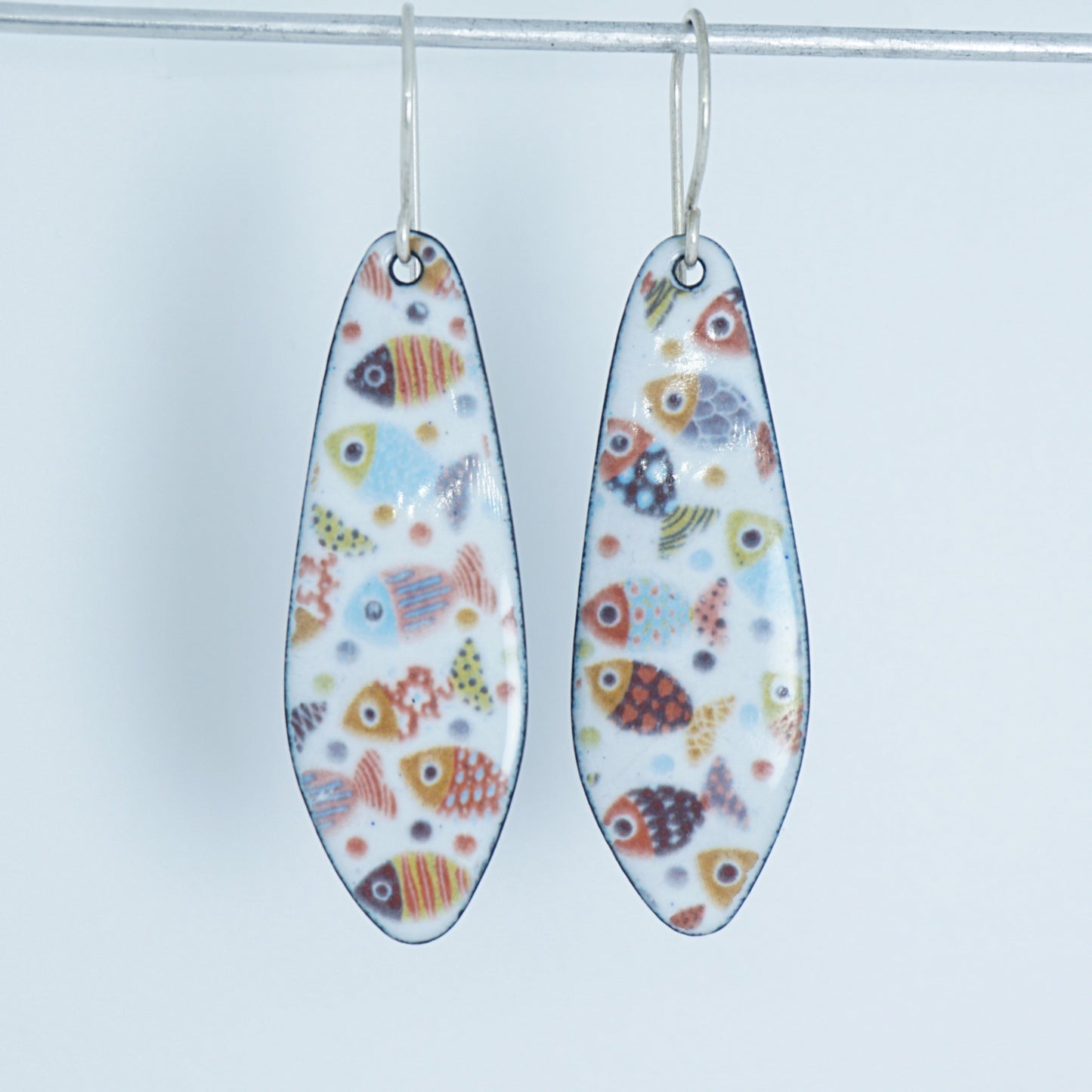 A pair of teardrop-shaped dangling earrings with a white background and a colorful pattern of small fish, hanging from silver hooks against a white background.