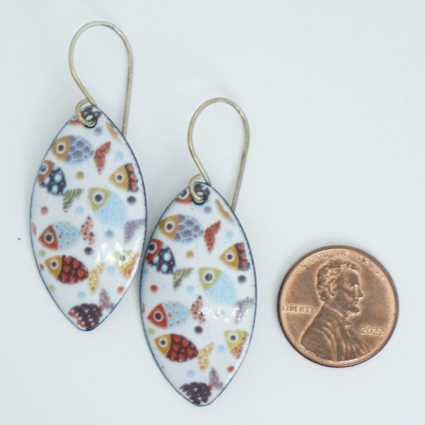 a pair of marquis shaped white enamel earrings with a colorful fish print, next to a penny for scale, against a white background