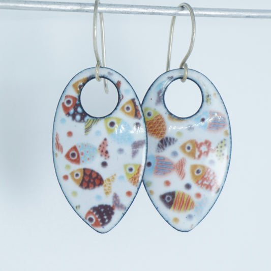 A pair of teardrop-shaped dangling earrings with a white enamel background decorated with colorful fish, suspended from silver wire hooks.