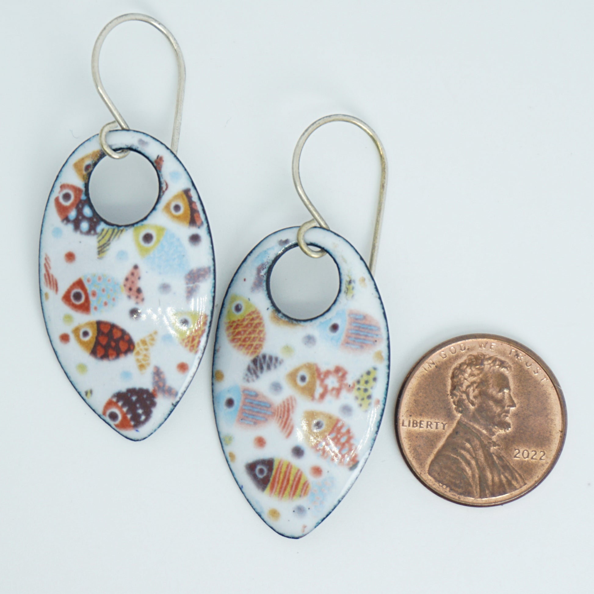 a pair of teardrop-shaped dangling earrings with a colorful fish pattern on a white background, shown next to a penny for scale