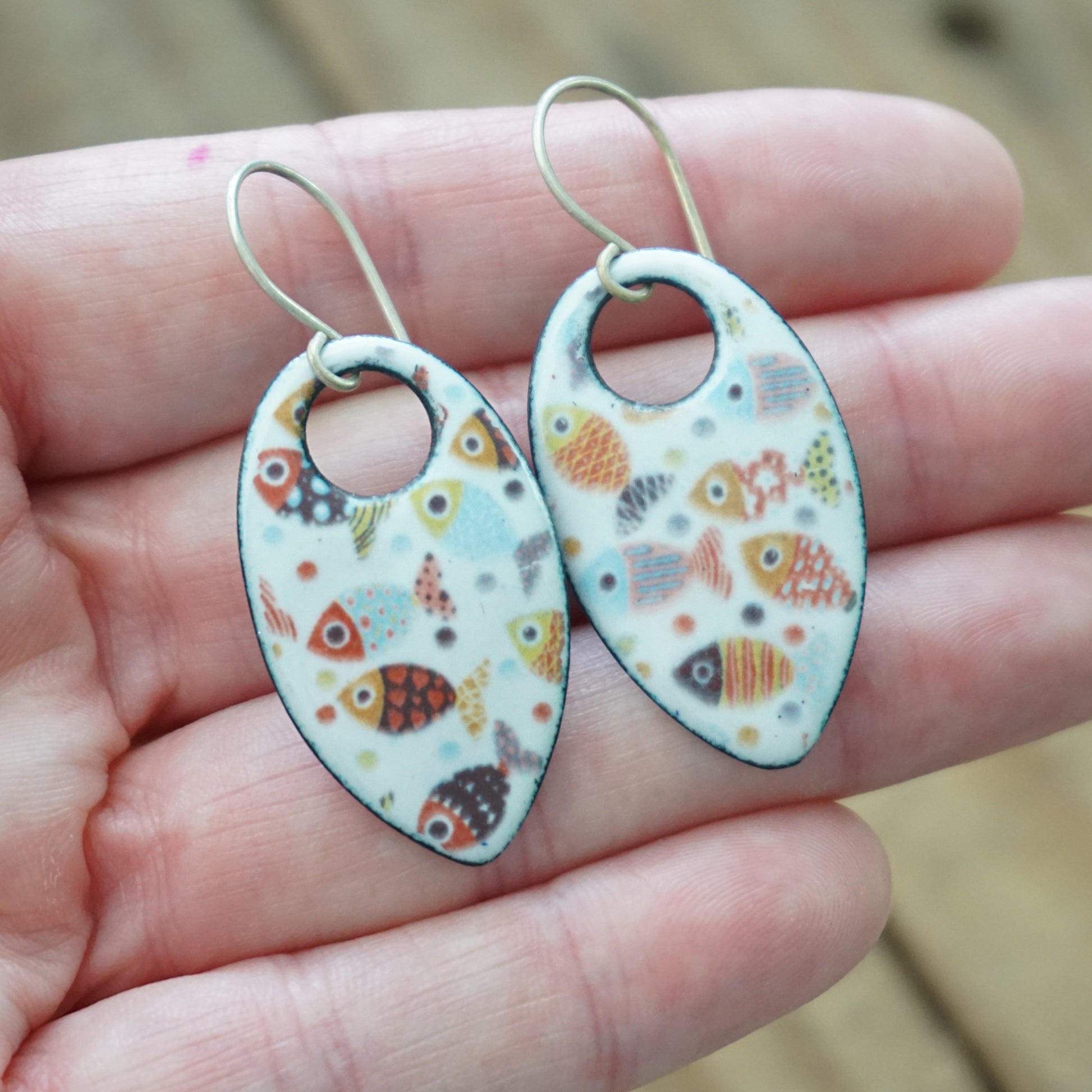 A pair of teardrop shaped fish earrings with a white enamel background and colorful fish patterns, held in a person's hand.