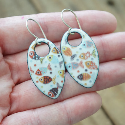A pair of teardrop shaped fish earrings with a white enamel background and colorful fish patterns, held in a person's hand.
