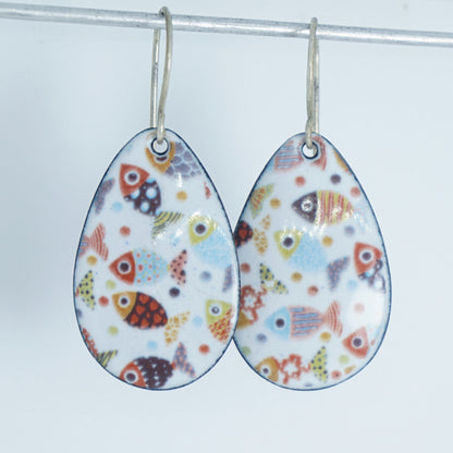a pair of teardrop shaped white enamel earrings with a colorful fish print, against a white background