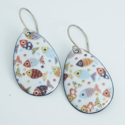 a pair of teardrop shaped white enamel earrings with a colorful fish print, against a white background
