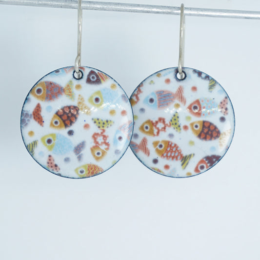 A pair of round white enamel dangle earrings decorated with a colorful pattern of stylized fish, hanging from silver hooks against a plain light background.