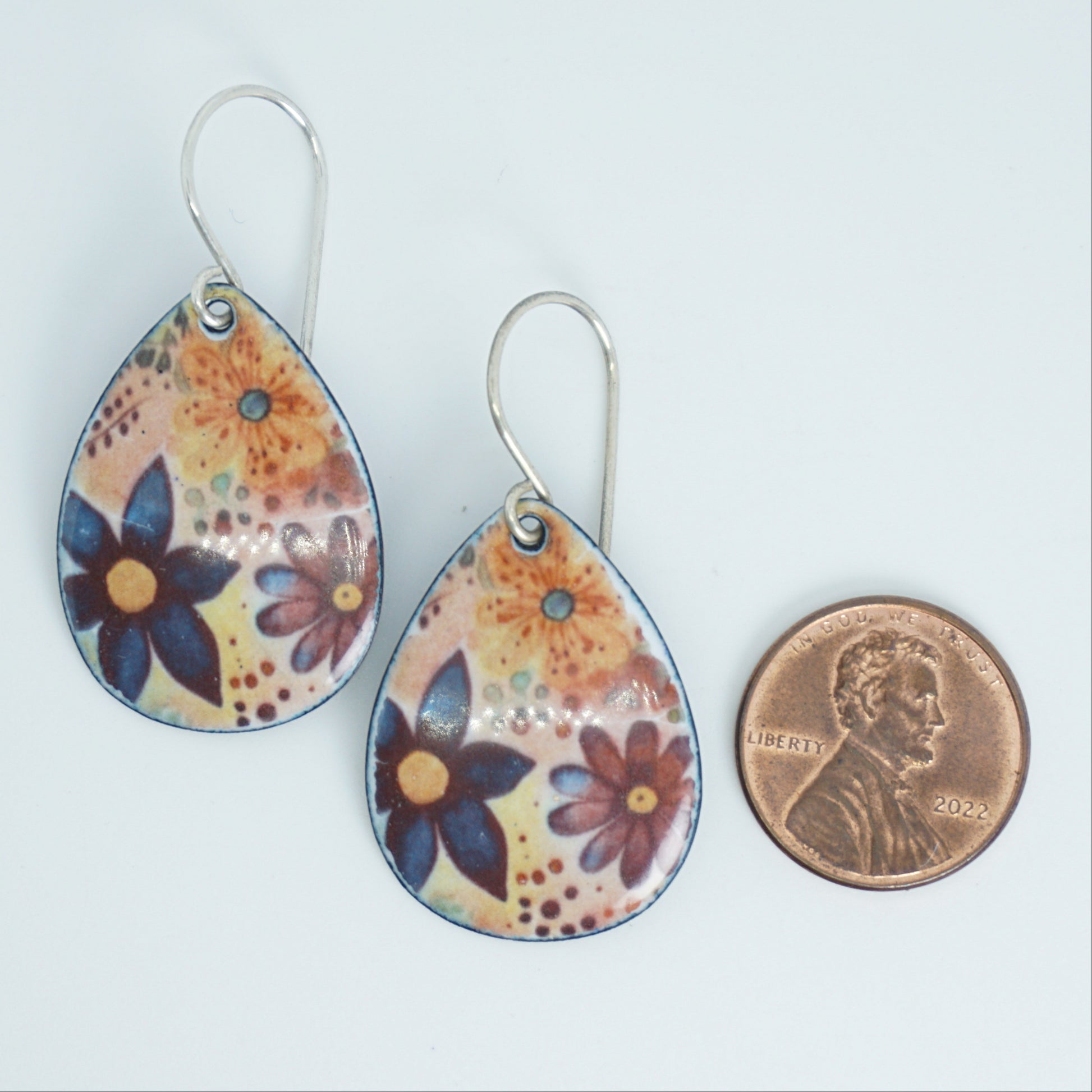 Teardrop-shaped dangling earrings with a colorful floral pattern on a light background, shown next to a penny for scale on a plain white surface.