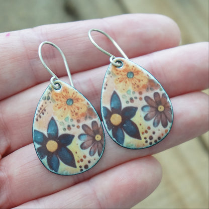 A pair of teardrop-shaped dangle earrings with a floral pattern, held in a person's hand. The earrings feature dark blue, orange, and brown flowers with a silver fishhook wire.