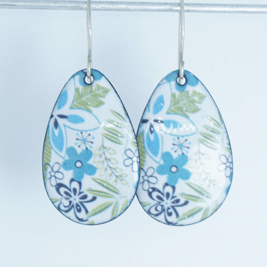 A pair of teardrop-shaped dangle earrings with a white enamel background decorated with blue flowers, green leaves, and dark blue floral accents, hanging from silver hooks.