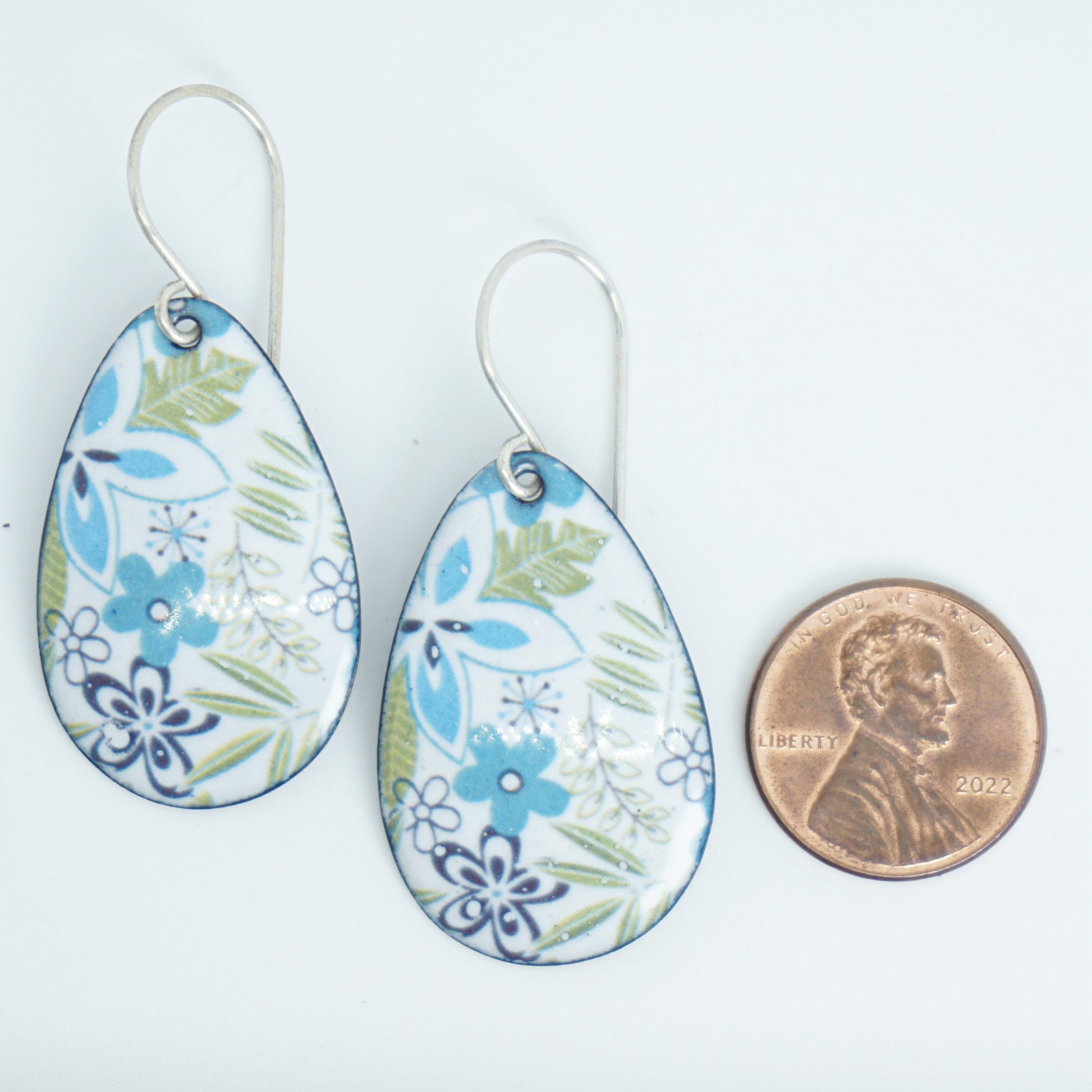 Teardrop-shaped dangle earrings with a blue, green, and white floral pattern, shown next to a US penny for scale on a white background.