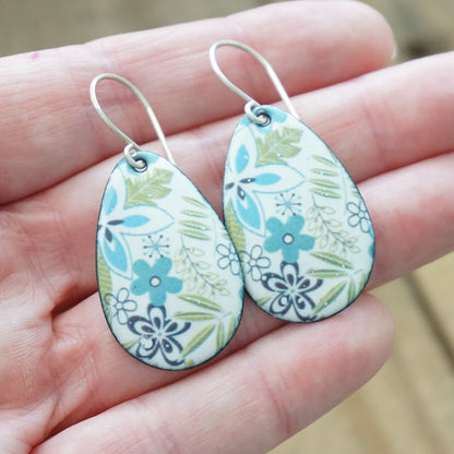 A pair of teardrop-shaped dangle earrings with a floral pattern, held in a person's hand. The earrings feature a white background with blue, green, and purple floral motifs, suspended from silver fishhook wires.