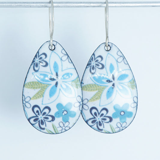 A pair of teardrop-shaped dangling earrings with a white background and a floral pattern in light blue, purple, and light green, hanging from silver hooks against a white background.