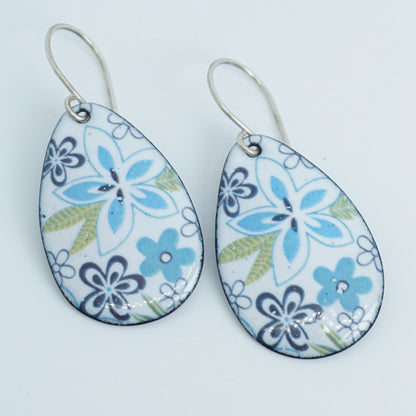 A pair of teardrop-shaped dangling earrings with a white enamel background, decorated with blue and purple floral motifs and light green leaves, suspended from silver fishhook wires.
