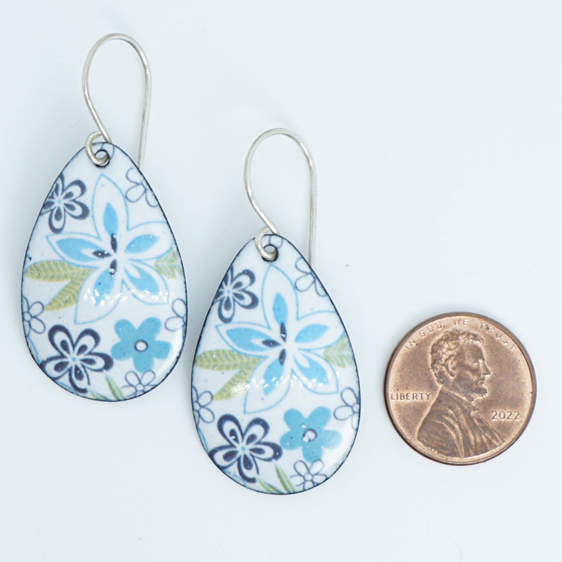 A pair of teardrop-shaped dangle earrings with a white background, blue and purple floral pattern, and light green leaves, shown next to a US penny for scale on a white background