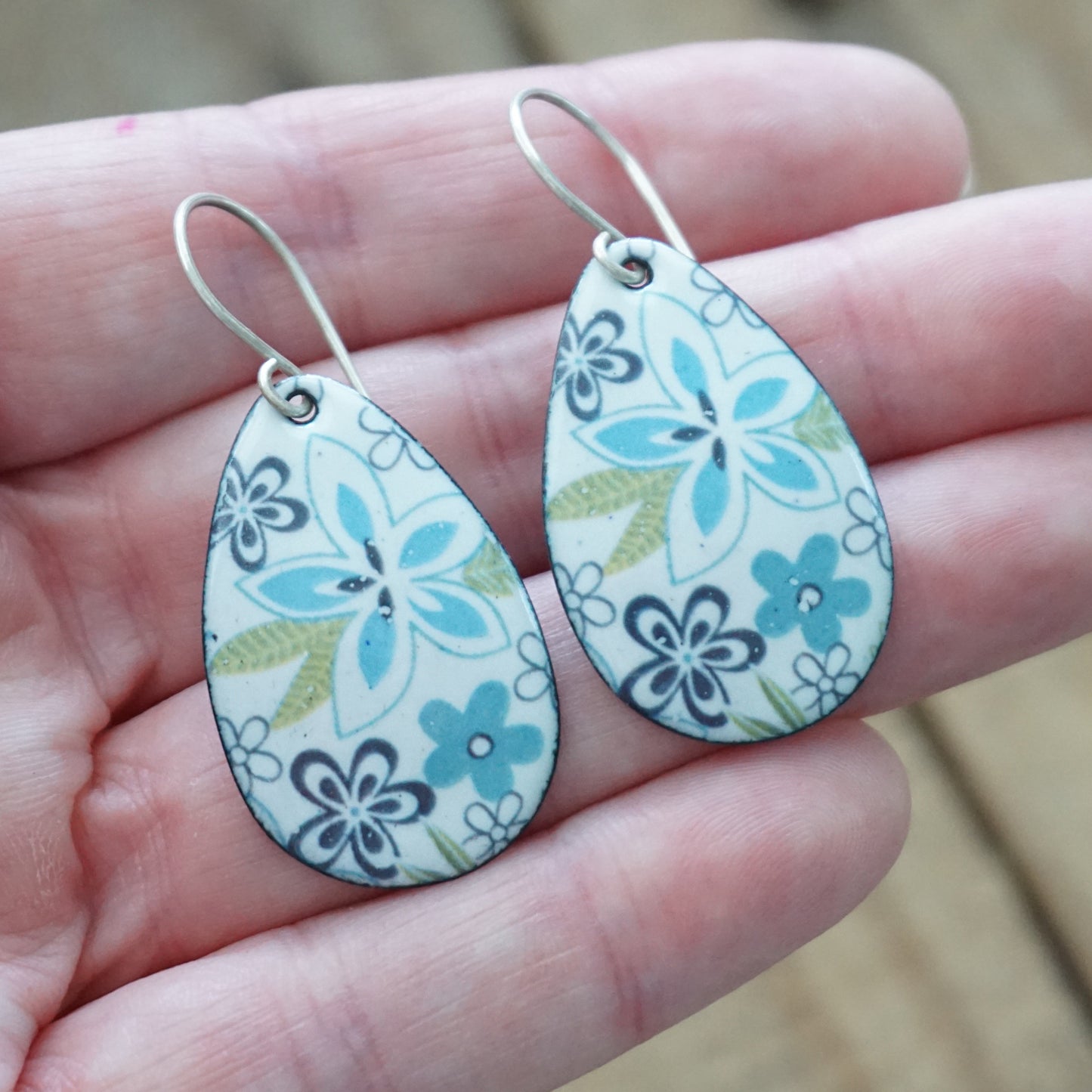 Teardrop-shaped earrings with a floral pattern in light blue, purple, and olive green on a white background, held in a person's hand.