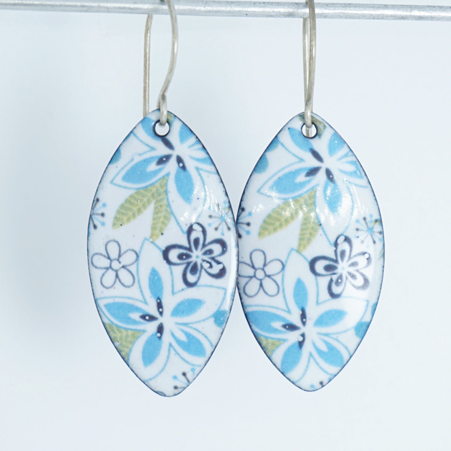 A pair of dangling marquise-shaped earrings with a white background and a purple, light blue, and green floral pattern, hanging from thin silver hooks against a plain white background.