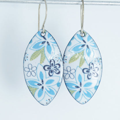 A pair of dangling marquise-shaped earrings with a white background and a purple, light blue, and green floral pattern, hanging from thin silver hooks against a plain white background.