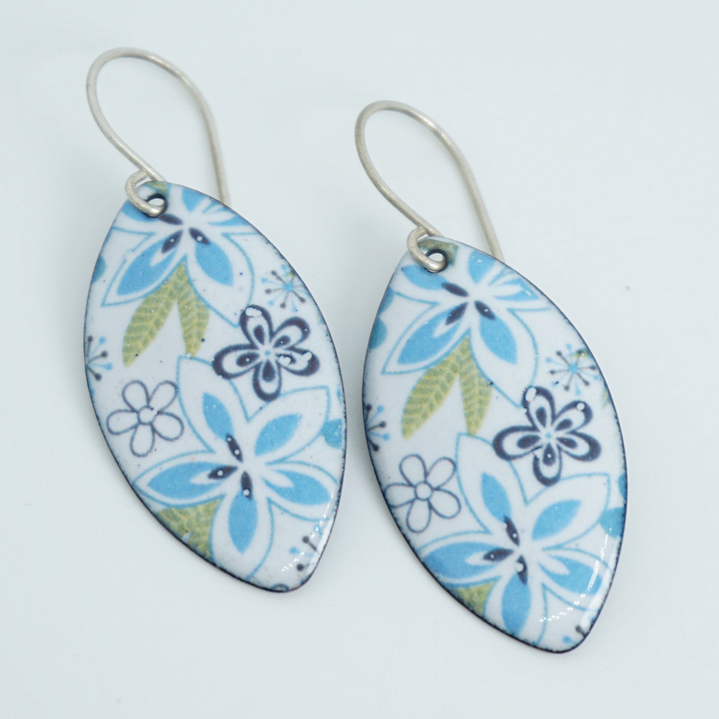 A pair of marquis-shaped drop earrings with a white enamel background decorated with blue and green floral patterns, suspended from silver-tone wire hooks.