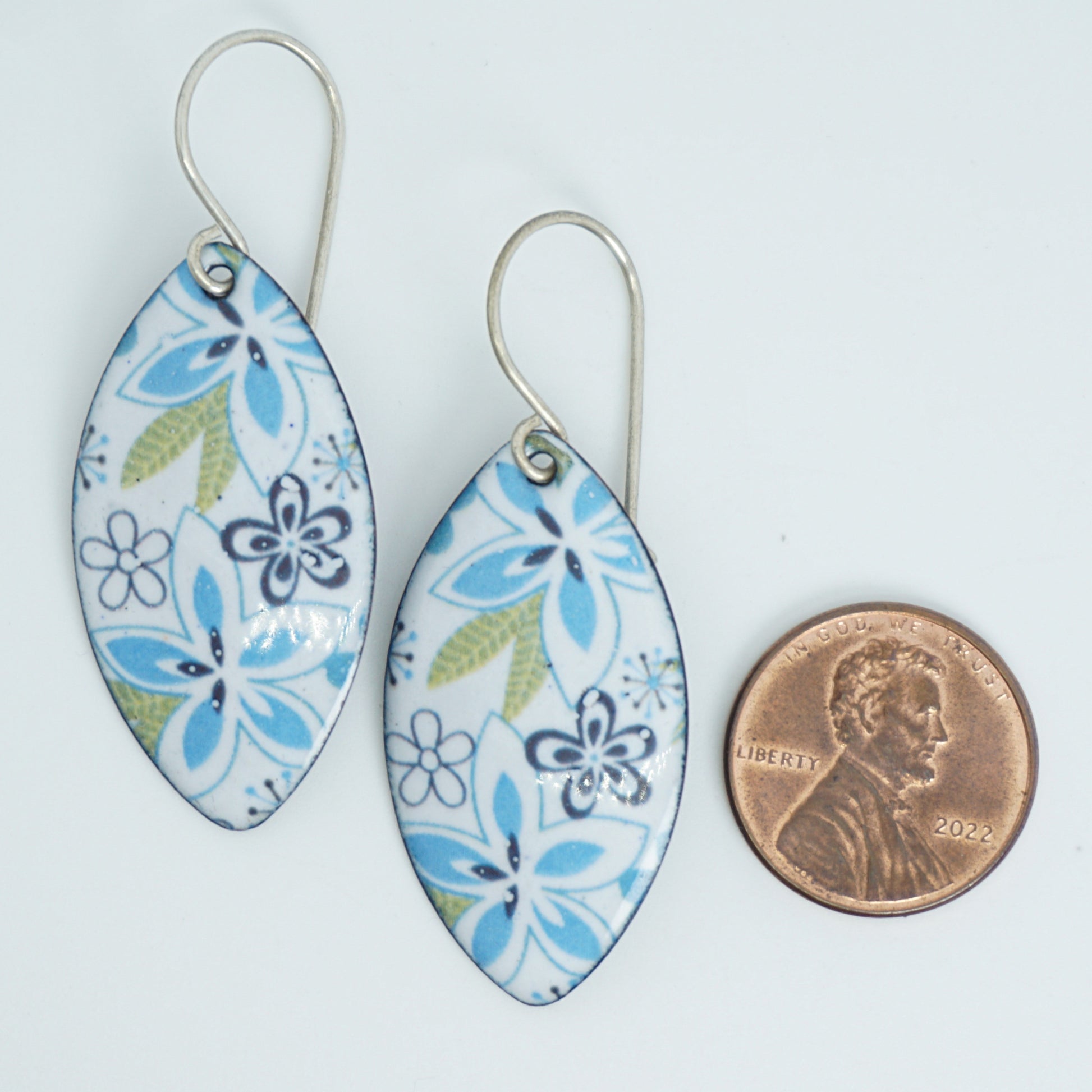 A pair of dangling marquise-shaped earrings with a white background and a blue, green, and purple floral pattern, shown next to a penny for scale, against a white background