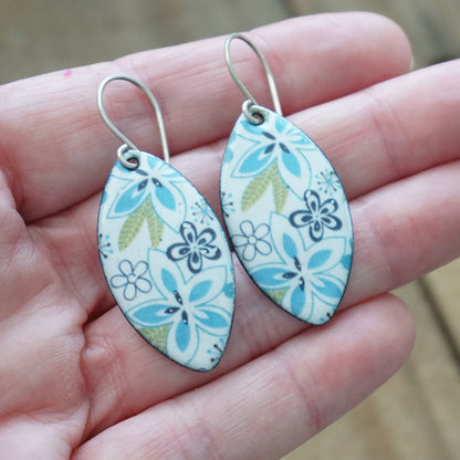 A pair of marquis shaped earrings with floral designs in light blue, purple, and olive green on a white background, held in a person's hand.