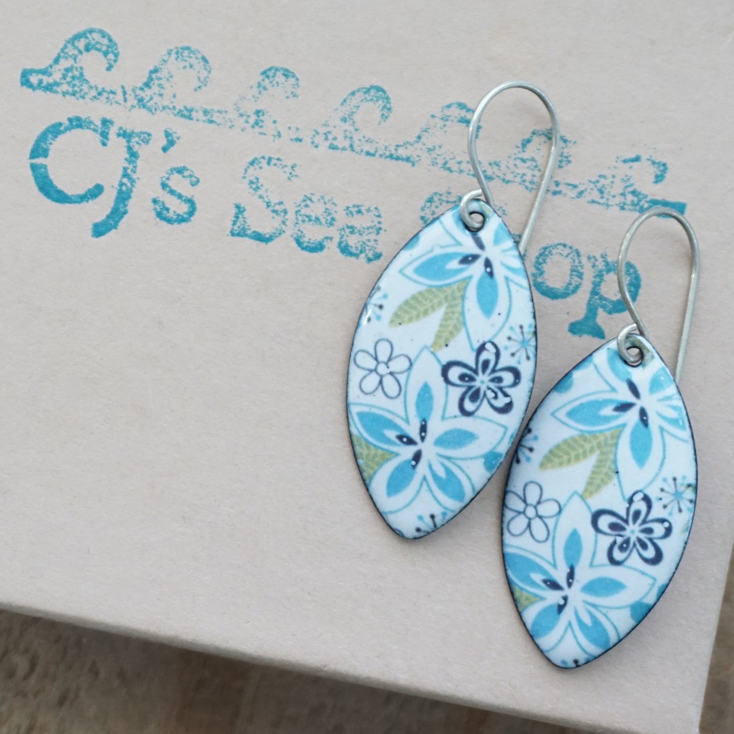A pair of marquis-shaped dangle earrings with a light blue and white floral pattern, displayed on a gift box