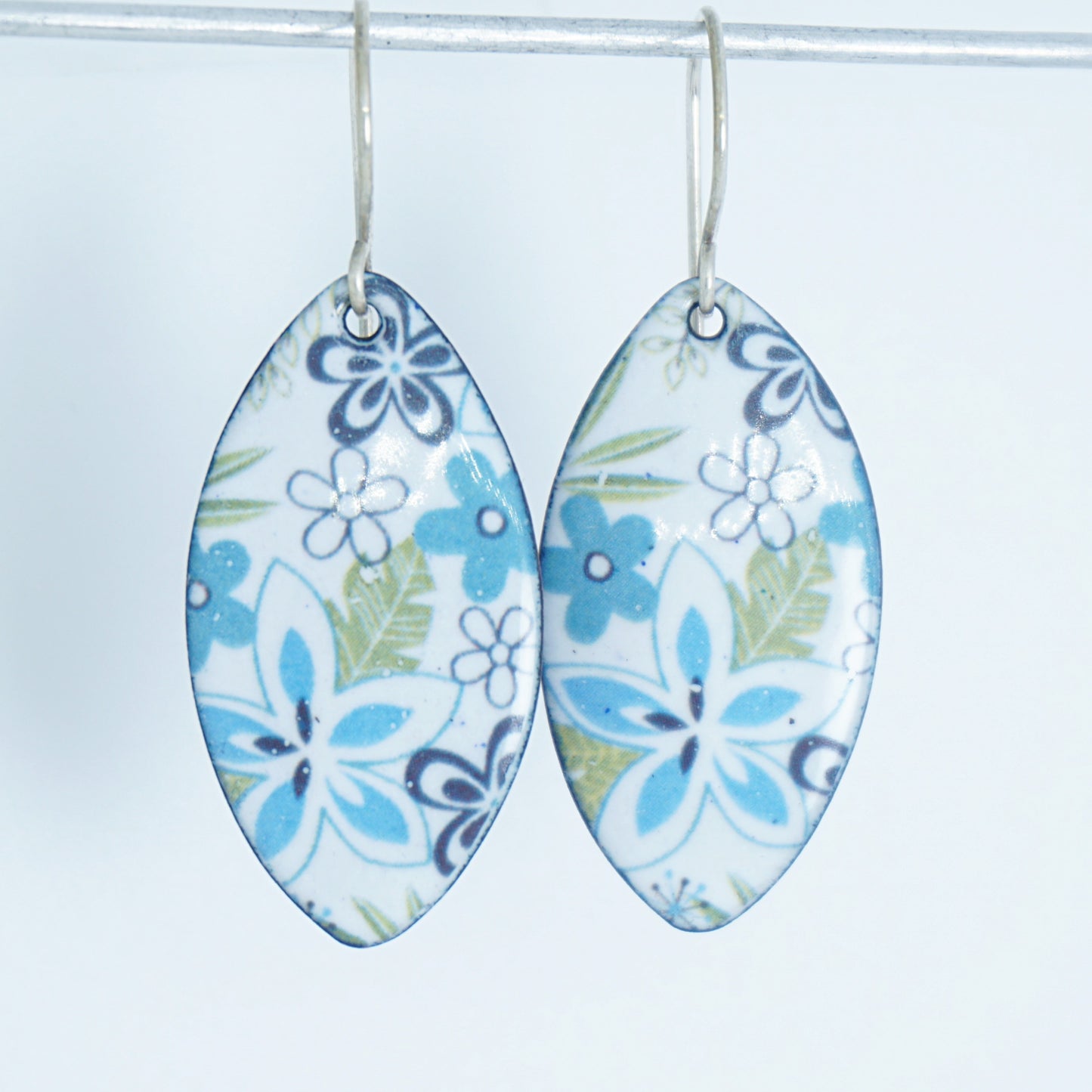 A pair of dangling marquise-shaped earrings with a white background and a purple, light blue, and green floral pattern, hanging from silver hooks against a white background.