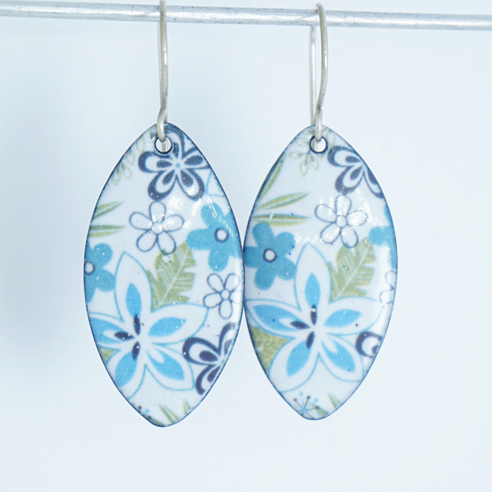 A pair of dangling marquise-shaped earrings with a white background and a purple, light blue, and green floral pattern, hanging from silver hooks against a white background.