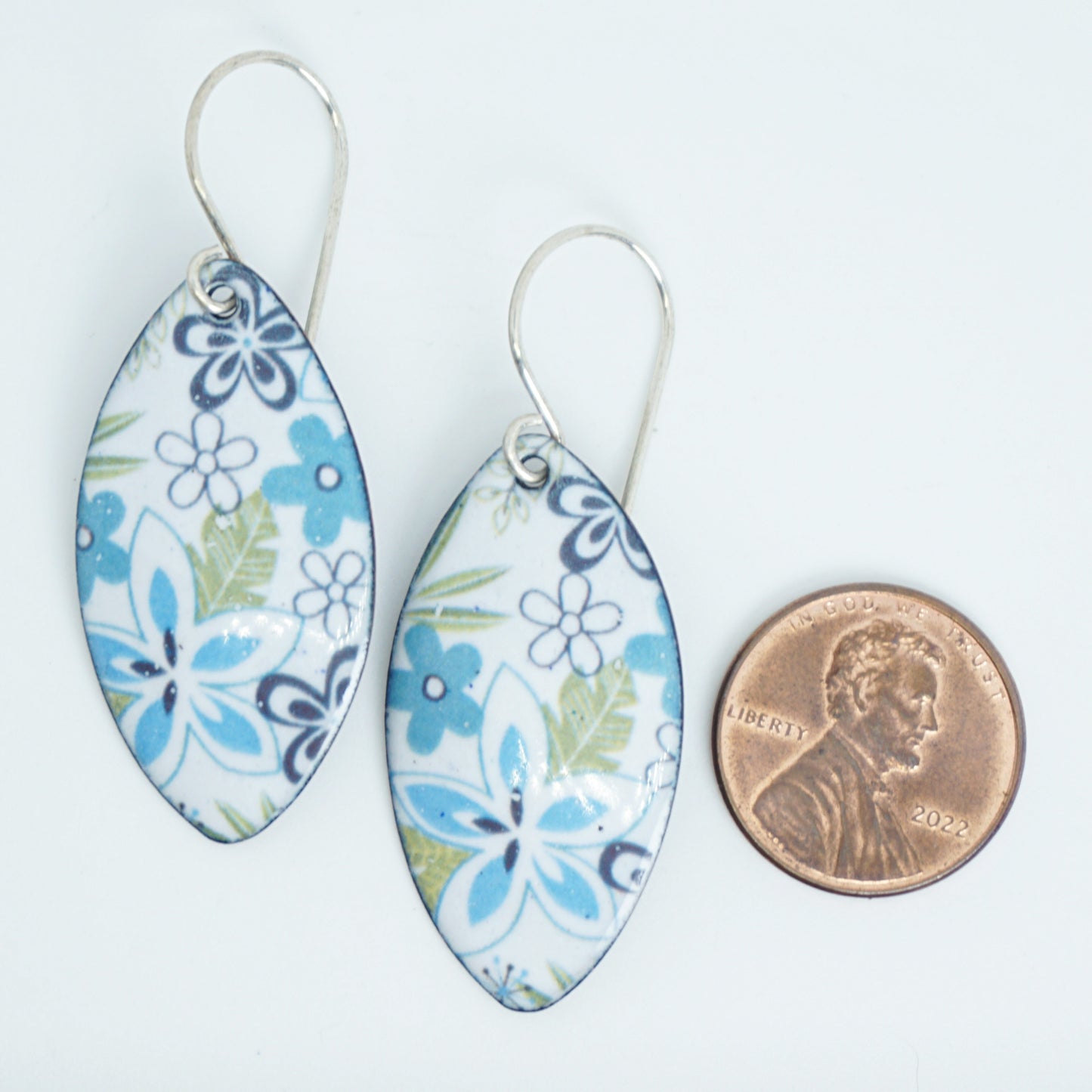 A pair of marquise-shaped white enamel earrings decorated with blue and green floral patterns, shown next to a US penny for scale on a white background.