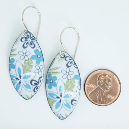 A pair of marquise-shaped white enamel earrings decorated with blue and green floral patterns, shown next to a US penny for scale on a white background.
