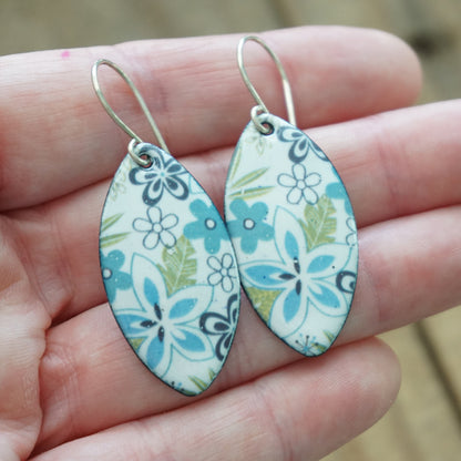 A pair of handmade marquis-shaped dangle earrings with a blue, green, and purple floral pattern on a white background, held in a person's hand