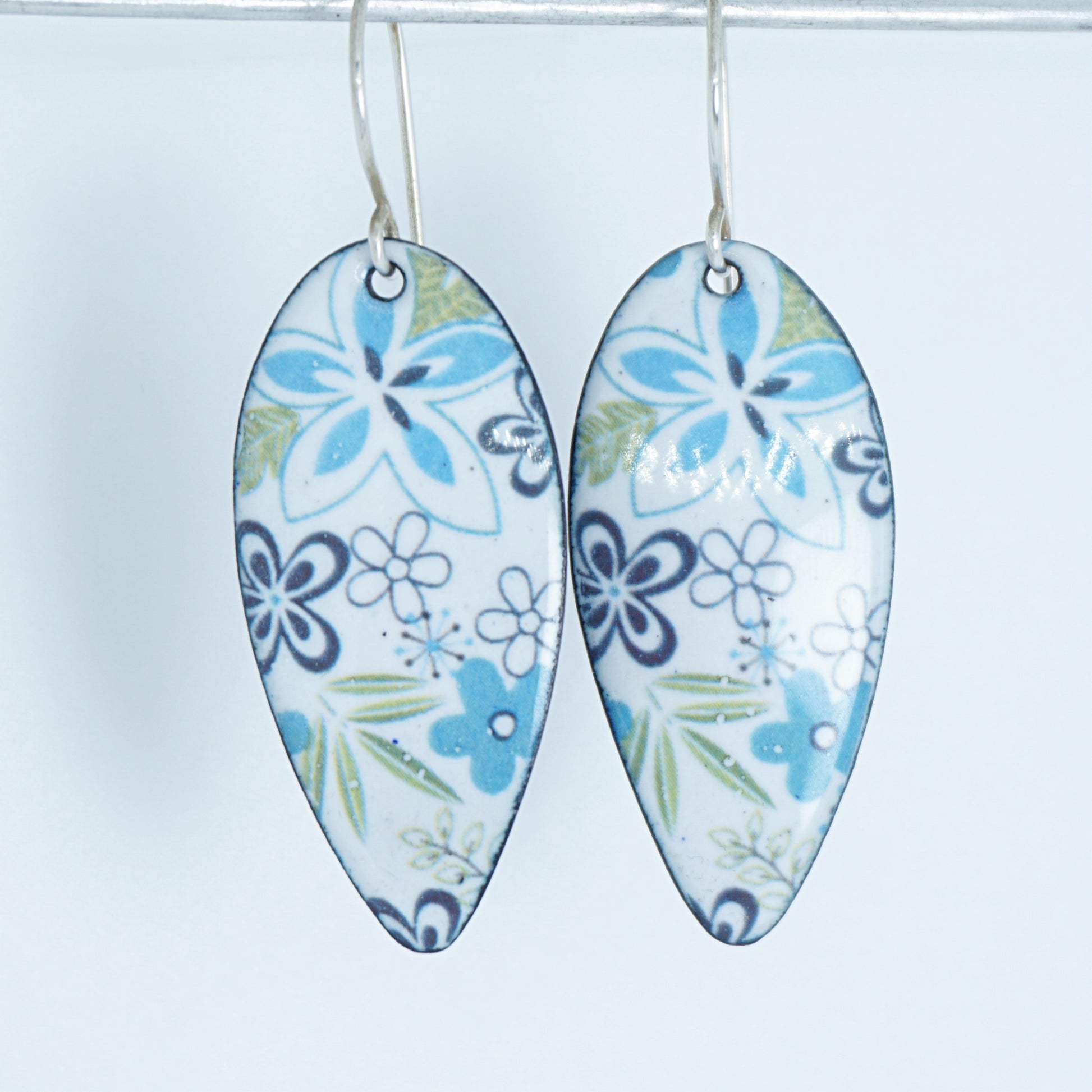 A pair of teardrop-shaped white enamel pendants decorated with a blue, purple, and green floral pattern, hanging against a plain white background.