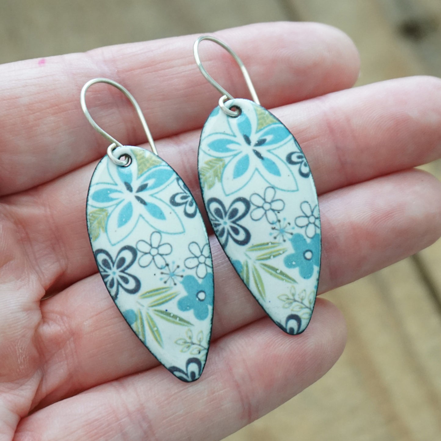 A pair of teardrop-shaped dangle earrings with a blue, green, and purple floral pattern on a white background, held in a person's hand.