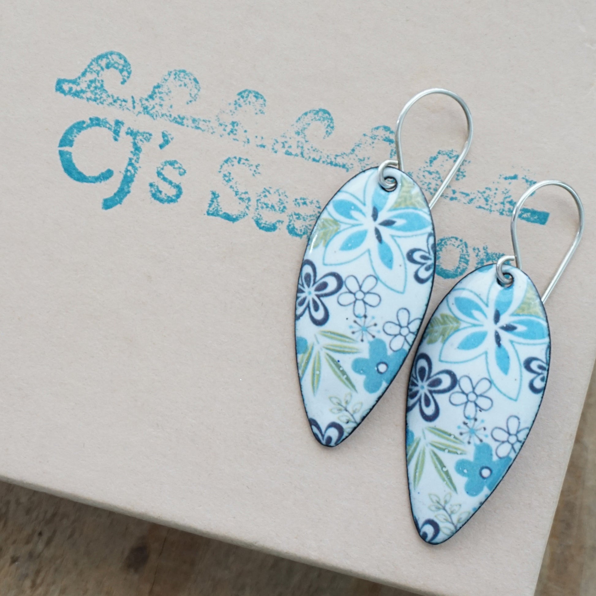 A pair of teardrop-shaped dangle earrings with a blue and white floral pattern, displayed on a gift box