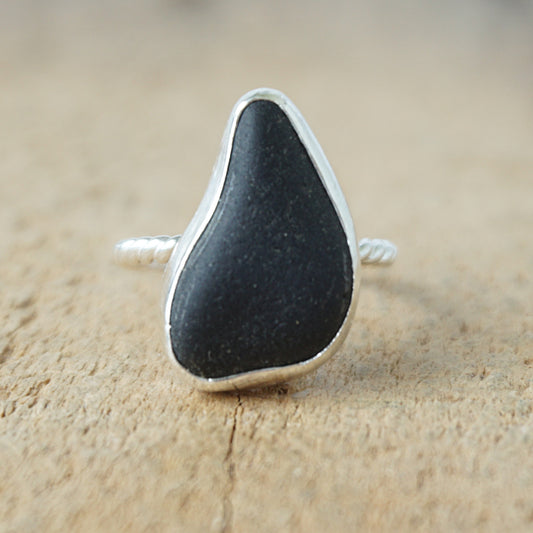 A sterling silver ring featuring teardrop-shaped black sea glass, on a textured light brown wood surface.