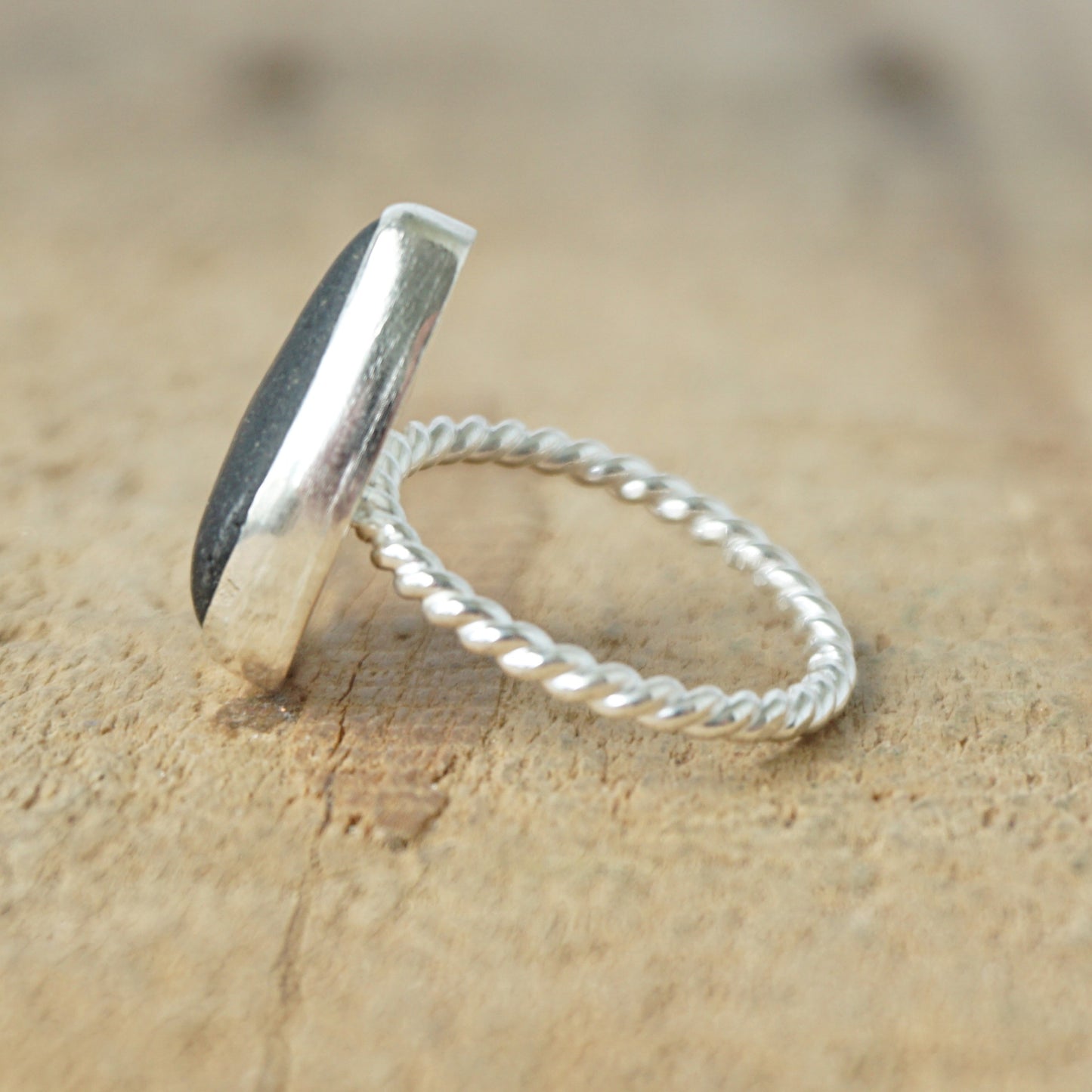 side view of A sterling silver ring featuring teardrop-shaped black sea glass, on a textured light brown wood surface.