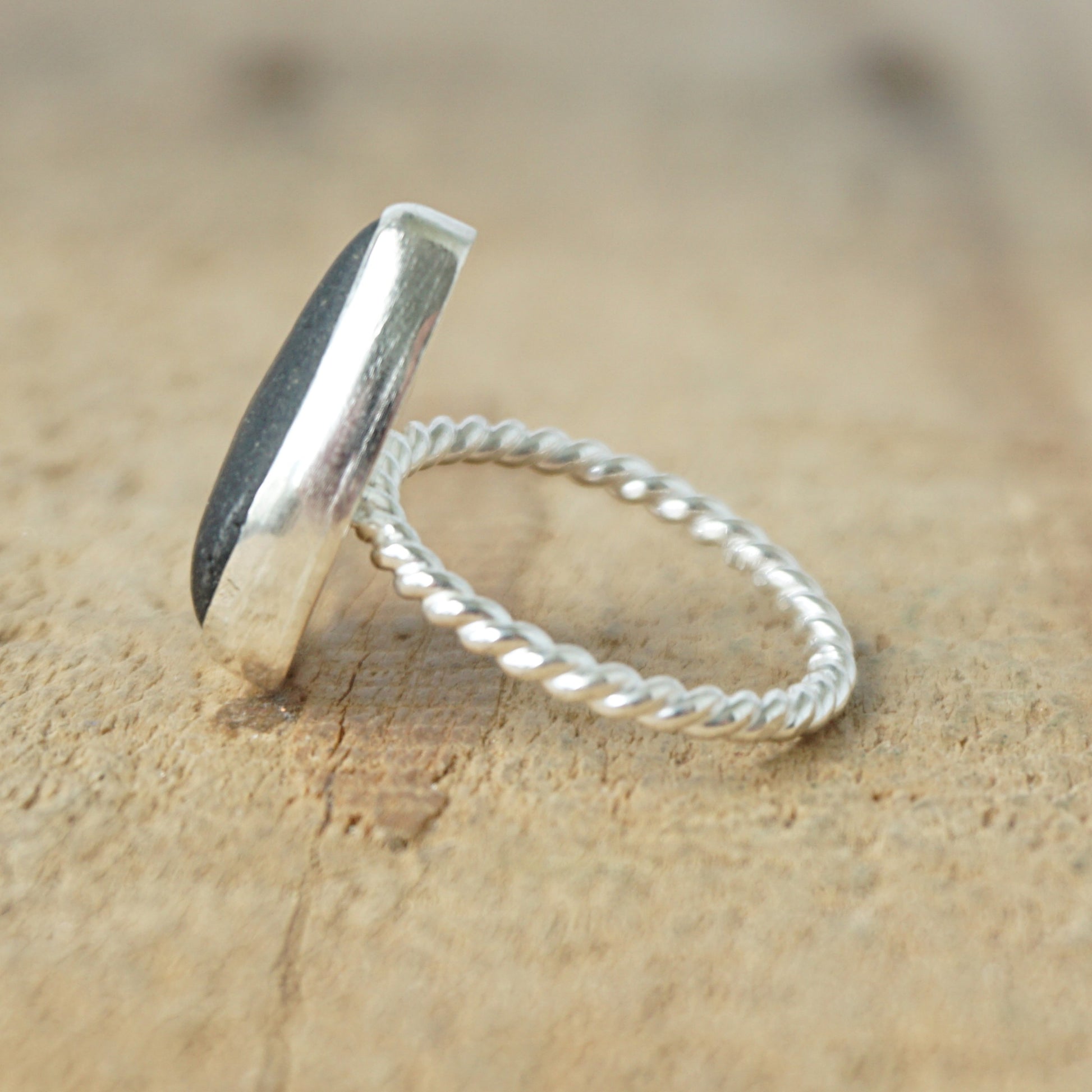 side view of A sterling silver ring featuring teardrop-shaped black sea glass, on a textured light brown wood surface.