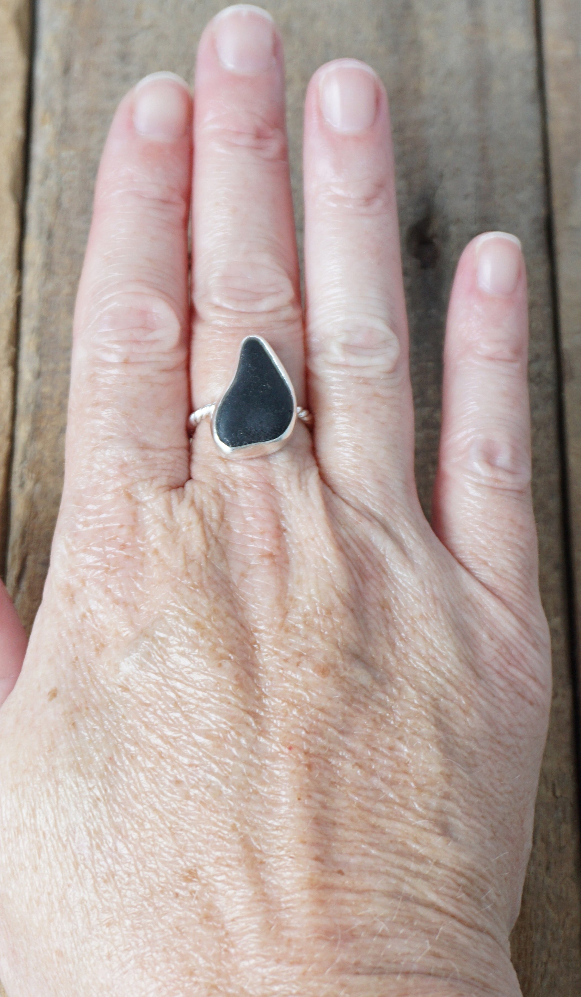 hand wearing A sterling silver ring featuring teardrop-shaped black sea glass, on a textured light brown wood surface.