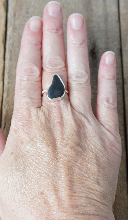 hand wearing A sterling silver ring featuring teardrop-shaped black sea glass, on a textured light brown wood surface.