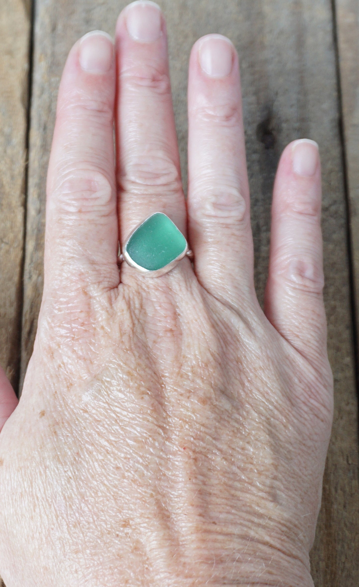 hand wearing a silver ring with dark green sea glass, on a wooden surface.