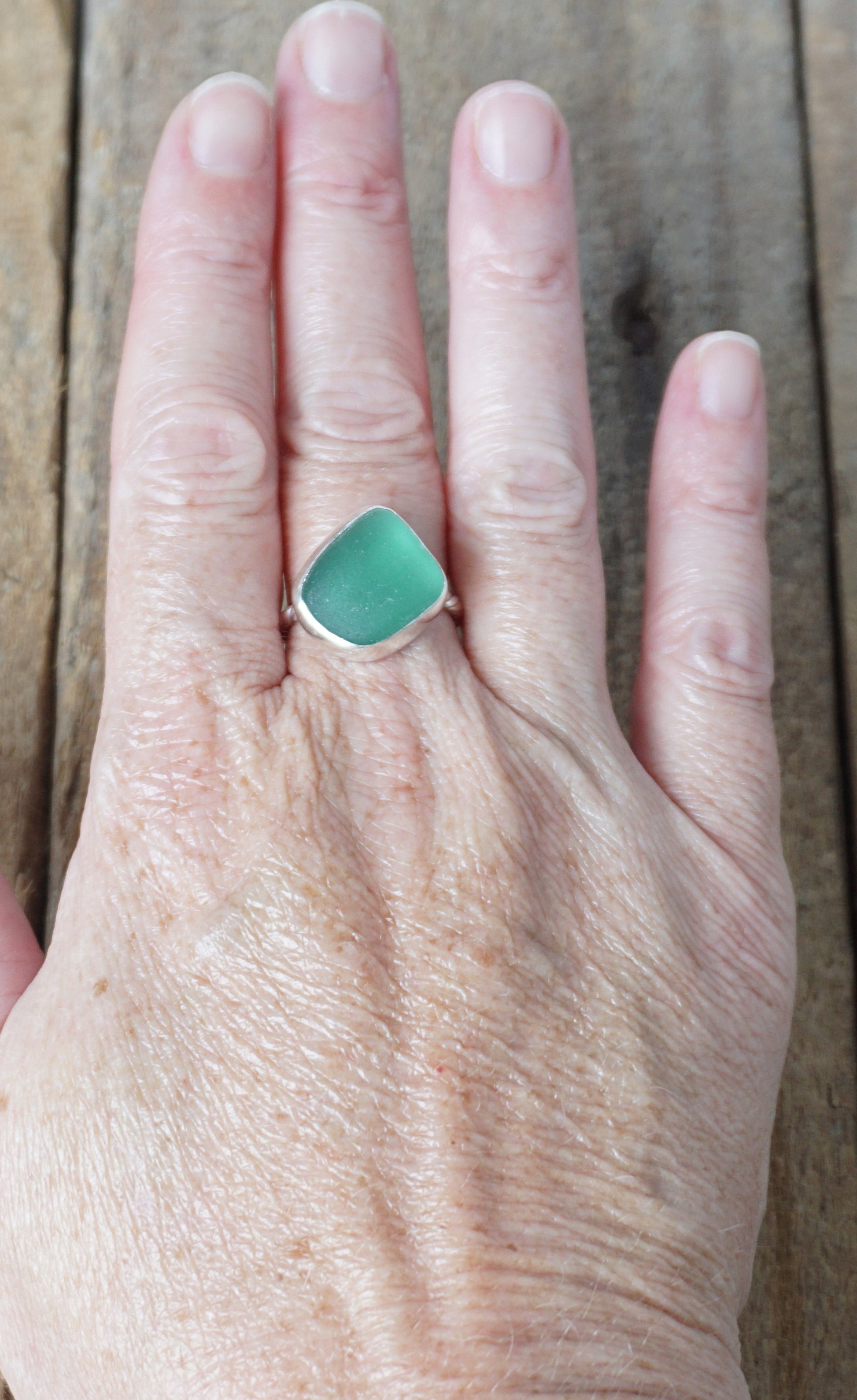 hand wearing a silver ring with dark green sea glass, on a wooden surface.