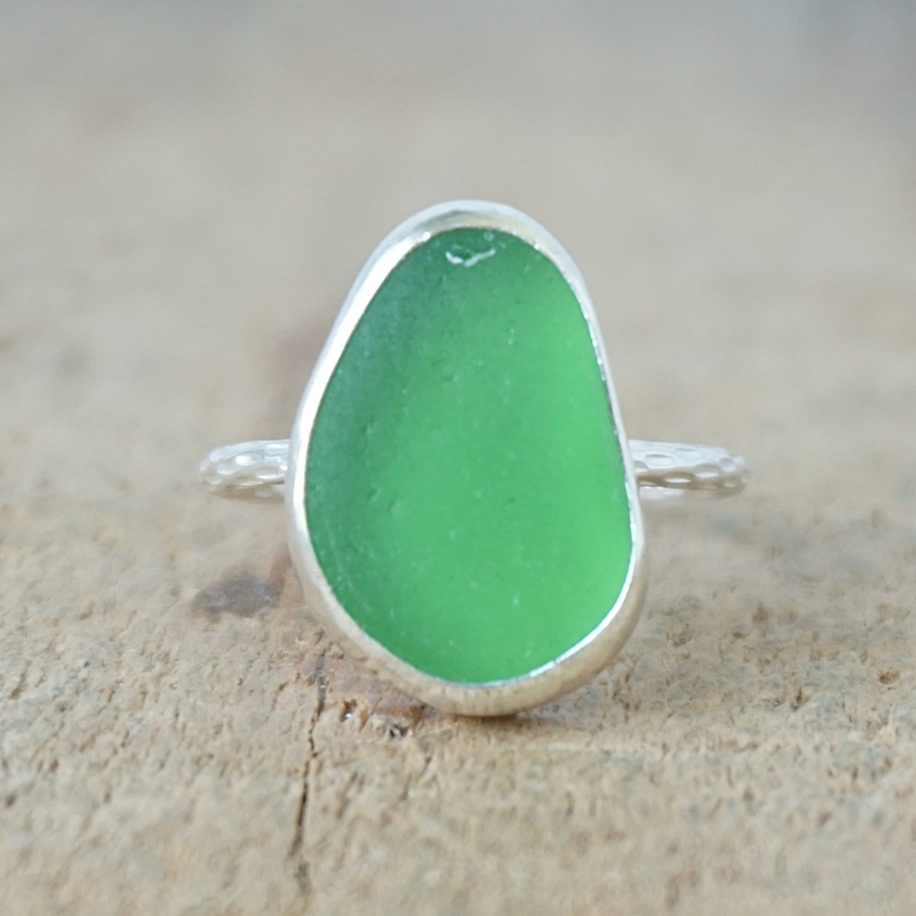 A sea glass ring featuring a vibrant green teardrop-shaped piece of frosted sea glass set in a silver bezel, resting on a textured beige surface.