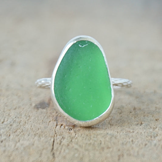 A sea glass ring featuring a vibrant green teardrop-shaped piece of frosted sea glass set in a silver bezel, resting on a textured beige surface.