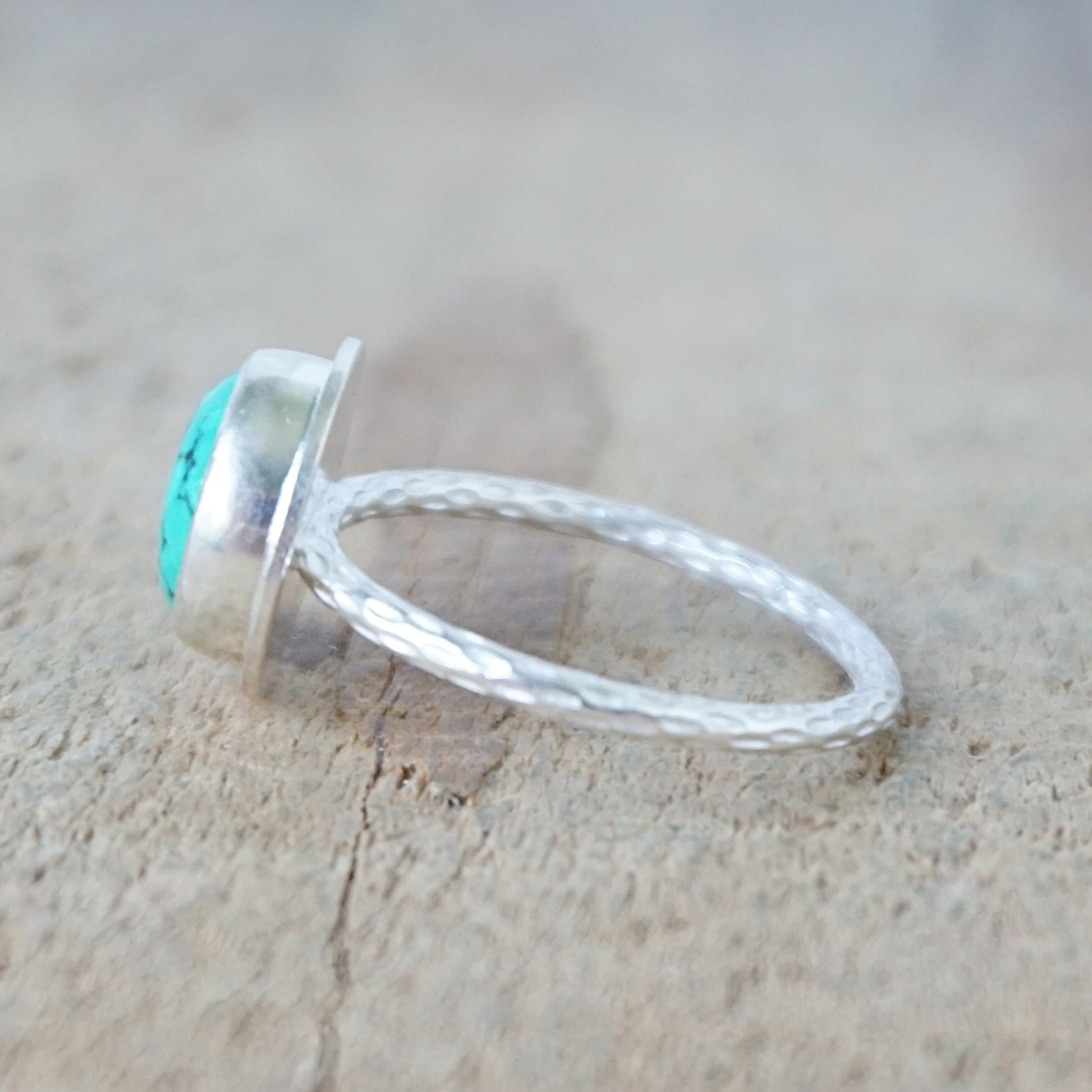 side view of A sterling silver ring featuring a round turquoise cabochon stone with natural dark veining, set against a neutral, textured beige background.