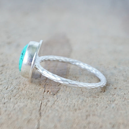 side view of A sterling silver ring featuring a round turquoise cabochon stone with natural dark veining, set against a neutral, textured beige background.