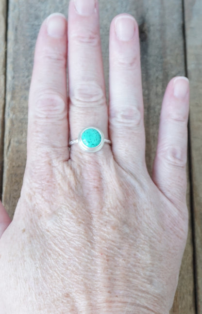 A silver ring with a round turquoise-colored stone, worn on a person's finger against a rustic wooden background.