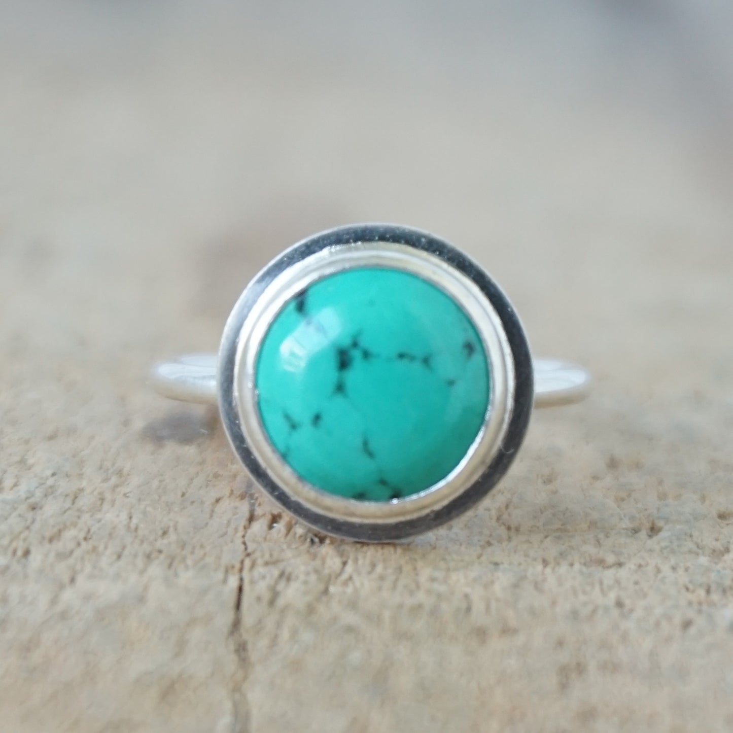 A sterling silver ring featuring a round turquoise cabochon stone with natural dark veining, set against a neutral, textured beige background.